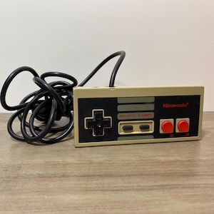 May include: A beige and black Nintendo controller with a black cable. The controller has a directional pad, select and start buttons, two red buttons, and the Nintendo logo.