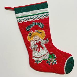 May include: Red quilted Christmas stocking with a green trim and a Cabbage Patch Kids design. The stocking features a Cabbage Patch Kid doll holding a candy cane and a gift.