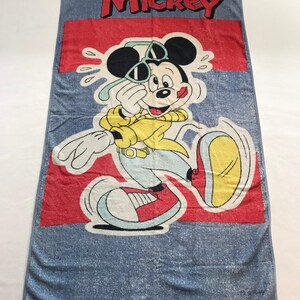 Pode incluir: Uma toalha de praia azul com uma faixa vermelha e uma imagem de desenho animado do Mickey Mouse usando &oacute;culos de sol e uma camisa amarela. O texto "Mickey" &eacute; impresso em vermelho no topo da toalha.