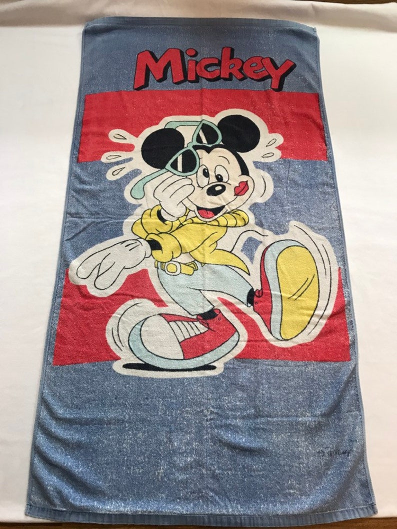 Pode incluir: Uma toalha de praia azul com uma faixa vermelha e uma imagem de desenho animado do Mickey Mouse usando &oacute;culos de sol e uma camisa amarela. O texto "Mickey" &eacute; impresso em vermelho no topo da toalha.