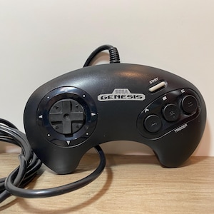 May include: Black Sega Genesis controller with a directional pad, start button, and A, B, and C buttons. The controller has the Sega Genesis logo and a trigger button. The controller is connected to a black cable.