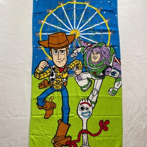 Puede incluir: Una toalla de playa verde y azul con una noria en el fondo. Los personajes Woody, Buzz Lightyear y Forky de la franquicia de pel&iacute;culas Toy Story aparecen en la toalla.