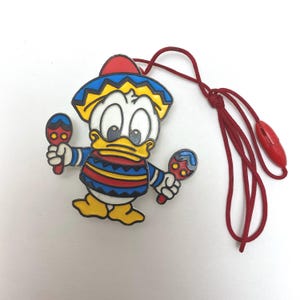 May include: A colorful plastic pendant of a cartoon duck wearing a sombrero and holding maracas. The pendant is attached to a red cord.