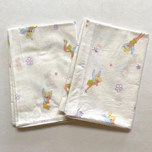 May include: Two folded white bed sheets featuring a repeating pattern of a cartoon fairy with blonde hair, green dress, and blue wings. The sheets also have small purple flowers and pink stars scattered across the surface.