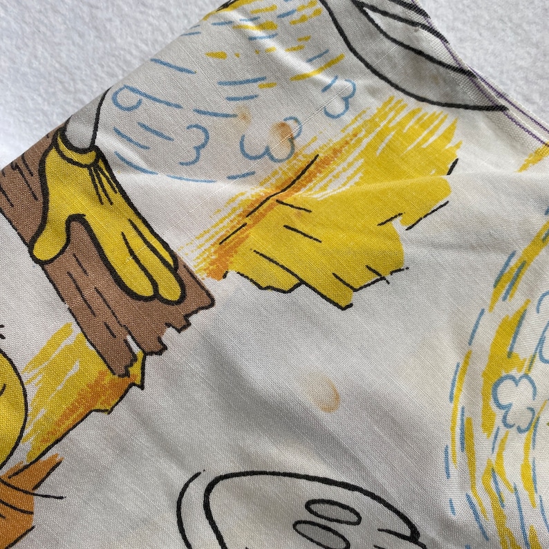 May include: A white fabric with a yellow, brown, and blue cartoon print. The print features a cartoon character wearing yellow gloves and holding a brown stick. The fabric has some brown stains.