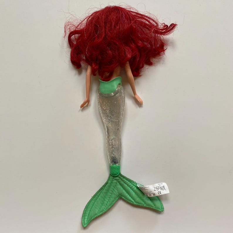 May include: A doll of a mermaid with red hair, a green top, and a silver tail. The mermaid's tail is green and has a tag attached. The doll is lying on a white surface.