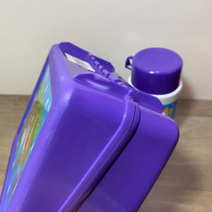 May include: Purple lunchbox with a cartoon design and a matching purple thermos.