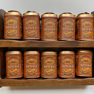 May include: A wooden spice rack with two shelves holds ten small, round, metal spice tins. The tins are all the same size and have a gold lid. Each tin has a label that reads "Famous Java Spices Imported".