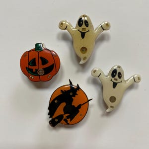 May include: Four Halloween themed pins. A white ghost with a smiling face, a pumpkin with a jack-o-lantern face, a witch flying on a broomstick, and another white ghost with a smiling face.