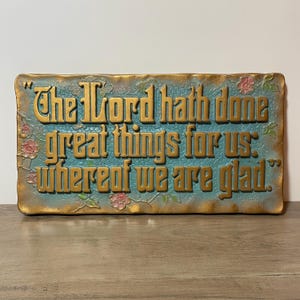 May include: Rectangular plaque with the text "The Lord hath done great things for us: whereof we are glad." in gold lettering. The plaque has a turquoise background with gold trim and pink floral accents.