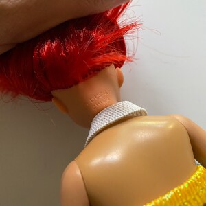 Puede incluir: Una mu&ntilde;eca de pelo rojo con un cuello blanco y una falda amarilla y naranja. La espalda de la mu&ntilde;eca es visible y el texto "DISNEY CHINA" est&aacute; impreso en la parte posterior del cuello de la mu&ntilde;eca.
