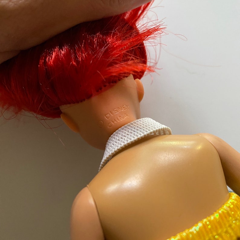 Puede incluir: Una mu&ntilde;eca de pelo rojo con un cuello blanco y una falda amarilla y naranja. La espalda de la mu&ntilde;eca es visible y el texto "DISNEY CHINA" est&aacute; impreso en la parte posterior del cuello de la mu&ntilde;eca.