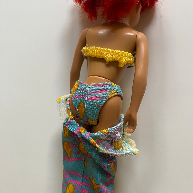 Puede incluir: Una mu&ntilde;eca con un traje de ba&ntilde;o azul y amarillo con estampado de peces. La mu&ntilde;eca lleva un pareo azul y amarillo con estampado de peces. La mu&ntilde;eca tiene el pelo rojo y lleva un collar amarillo.