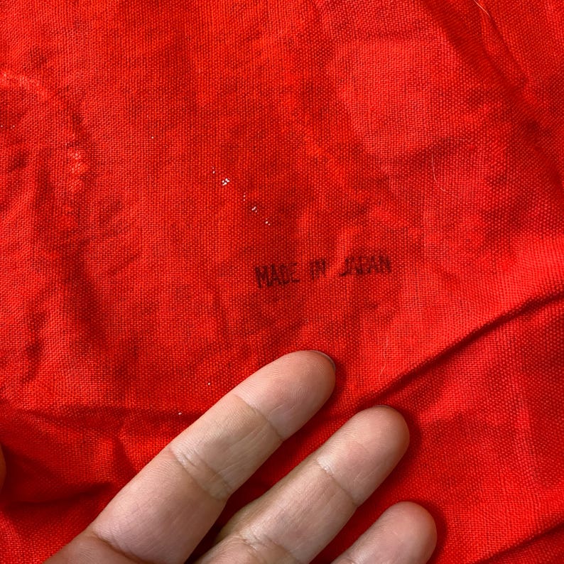 K&ouml;nnte beinhalten: Nahaufnahme eines leuchtend roten Stoffes mit dem Text "MADE IN JAPAN" in Schwarz gedruckt. Der Stoff hat eine strukturierte Optik und ist gefaltet, mit einer Hand, die teilweise im Bild zu sehen ist.
