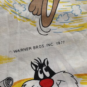May include: A white fabric with a cartoon image of Sylvester the Cat and Tweety Bird. The fabric has a yellow and blue background with clouds. The text "Warner Bros. Inc. 1977" is printed in black at the bottom of the fabric.