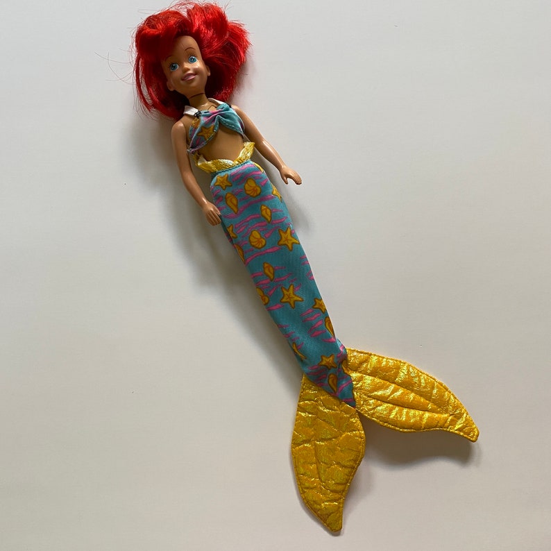 Puede incluir: Una mu&ntilde;eca sirena de pelo rojo con una cola azul y amarilla. La cola tiene un patr&oacute;n de conchas y estrellas de mar. La mu&ntilde;eca lleva un top de bikini rosa y blanco.