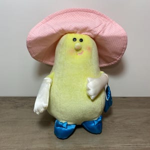 Peut inclure: Une peluche en forme de poire avec un chapeau rose à pois. Le corps vert clair a un visage souriant, des gants blancs et des chaussures bleues. Elle tient un petit sac à main bleu.