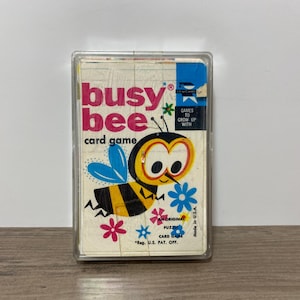 May include: A vintage Busy Bee card game deck in a clear plastic case. The box features a cartoon bee with yellow and black stripes, blue wings, and large eyes. Text includes 'busy bee card game', 'Games to grow up with', and 'Made in U.S.A.'