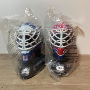 May include: Two miniature hockey goalie masks, one blue and white with the number 33, and the other red, white, and blue. Both are encased in clear plastic. The masks have a white cage and are likely collectibles or souvenirs.