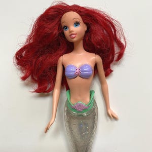 May include: A doll of a mermaid with long, red hair and blue eyes. The doll is wearing a purple seashell top and a green and silver tail. The doll's tail is silver with glitter.