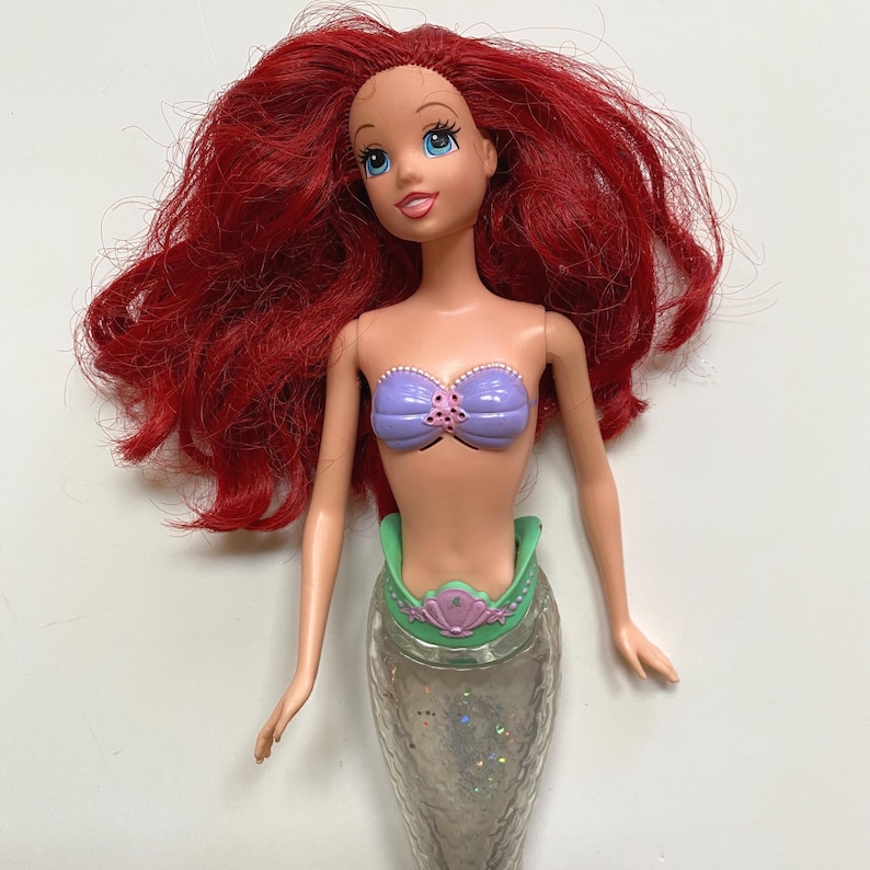 May include: A doll of a mermaid with long, red hair and blue eyes. The doll is wearing a purple seashell top and a green and silver tail. The doll's tail is silver with glitter.