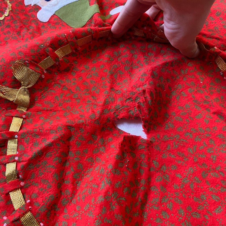 K&ouml;nnte beinhalten: Roter Weihnachtsbaumrock mit gr&uuml;nem Stechpalmenmuster. Der Rock hat eine goldene Borte und eine dekorative Schleife. Eine Hand h&auml;lt den Rand des Rocks.