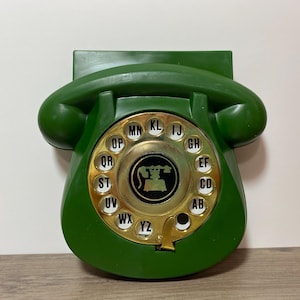 May include: A vintage green rotary dial telephone. The phone has a circular dial with letters and numbers, and a gold-colored center. The handset is curved and the phone is mounted on a wall.