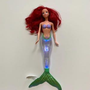 May include: A doll of a mermaid with red hair, a purple seashell top, and a light-up, clear tail. The mermaid's tail is green and has a fin. The doll is a toy.