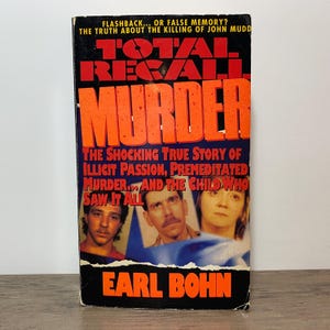 May include: A paperback book titled "Total Recall Murder" by Earl Bohn. The cover features the title in large orange letters over a blue background with three faces and a flag. The text reads "The shocking true story of illicit passion, premeditated murder... and the child who saw it all."