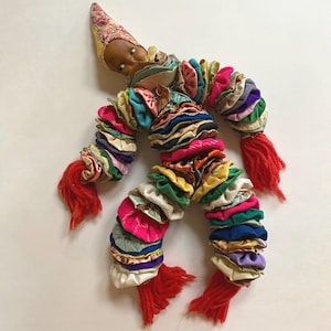 May include: A colourful handmade doll with a fabric body and a painted face. The doll's body is made of layered fabric circles in various colours, including pink, green, and yellow. Red yarn forms the doll's hands and feet, and a patterned hat tops the head.
