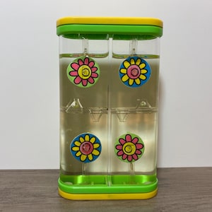 Peut inclure: Un minuteur liquide rectangulaire transparent avec un cadre jaune et vert. À l'intérieur, quatre décorations en forme de fleurs colorées flottent dans le liquide. Le minuteur est conçu pour mesurer le temps et a un aspect ludique.