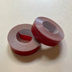 May include: Two rolls of red double-sided tape. The tape is wound around a cardboard core.