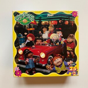 May include: A Cabbage Patch Kids 25-piece jigsaw puzzle. The puzzle box features a yellow border with the Cabbage Patch Kids logo and a scene of Cabbage Patch Kids in a red convertible in front of a diner.