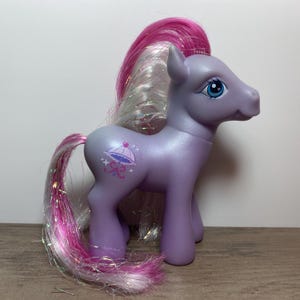 May include: A purple My Little Pony with a pink and white iridescent mane and tail. The pony has a blue eye and a symbol of a pink umbrella with stars on its flank.