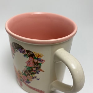 May include: Cream-colored ceramic mug with a pink interior and handle. The mug features a floral design with pink, purple, yellow, and brown flowers. The handle is a curved, off-white color.