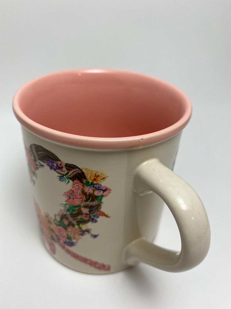 May include: Cream-colored ceramic mug with a pink interior and handle. The mug features a floral design with pink, purple, yellow, and brown flowers. The handle is a curved, off-white color.