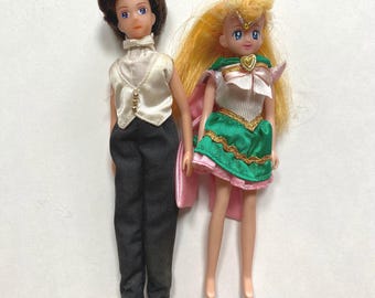 May include: Two small dolls, one with brown hair wearing a white shirt and black pants, and the other with blonde hair wearing a green and pink dress.