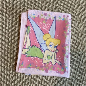 May include: A light pink pillowcase featuring a graphic illustration of a blonde fairy with blue eyes, wearing a green dress and translucent blue wings. The design includes a floral border with purple and green vines against a pink background.