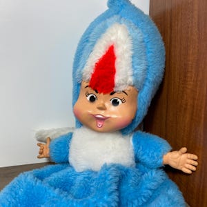 May include: A vintage doll in a blue, white, and red plush costume. The doll has a painted face with a smiling expression and outstretched arms. The doll's outfit includes a hood and a dress-like bottom.