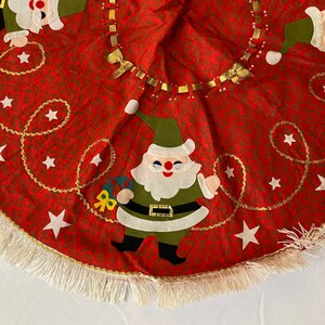 K&ouml;nnte beinhalten: Roter Weihnachtsbaumrock mit einem sich wiederholenden Muster aus kleinen gr&uuml;nen Punkten. Der Rock zeigt Applikationen von Weihnachtsmannfiguren, wei&szlig;en Sternen und goldfarbenen Verzierungen. Ein wei&szlig;er Fransenbesatz s&auml;umt den Rand des Rocks.