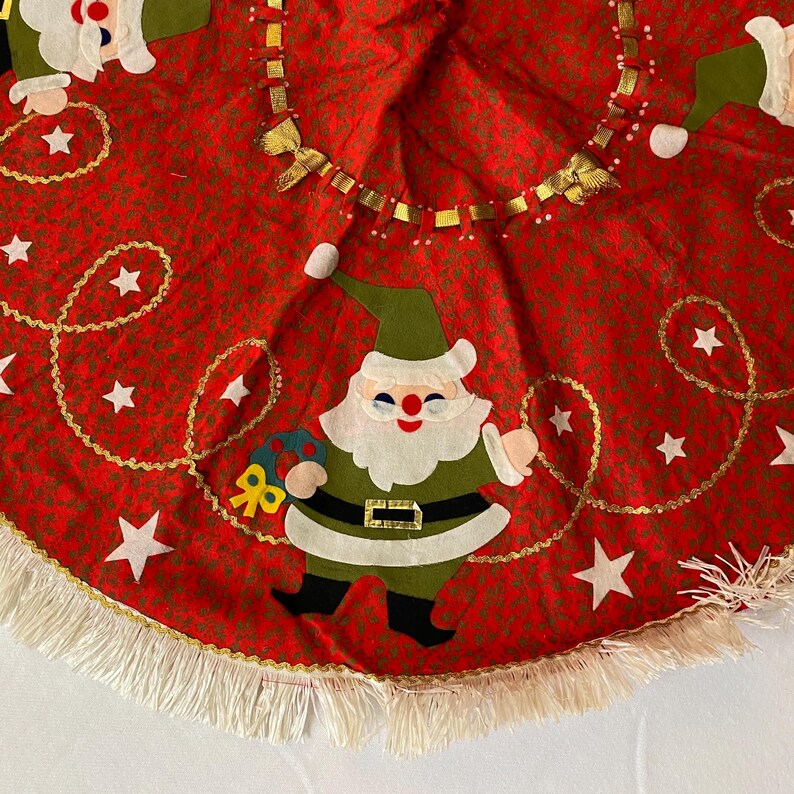 K&ouml;nnte beinhalten: Roter Weihnachtsbaumrock mit einem sich wiederholenden Muster aus kleinen gr&uuml;nen Punkten. Der Rock zeigt Applikationen von Weihnachtsmannfiguren, wei&szlig;en Sternen und goldfarbenen Verzierungen. Ein wei&szlig;er Fransenbesatz s&auml;umt den Rand des Rocks.