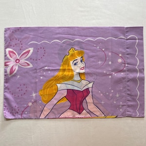 May include: A purple pillowcase with a white scalloped border features a cartoon image of Sleeping Beauty in her pink and yellow dress. The background is a light purple with white stars and swirls.