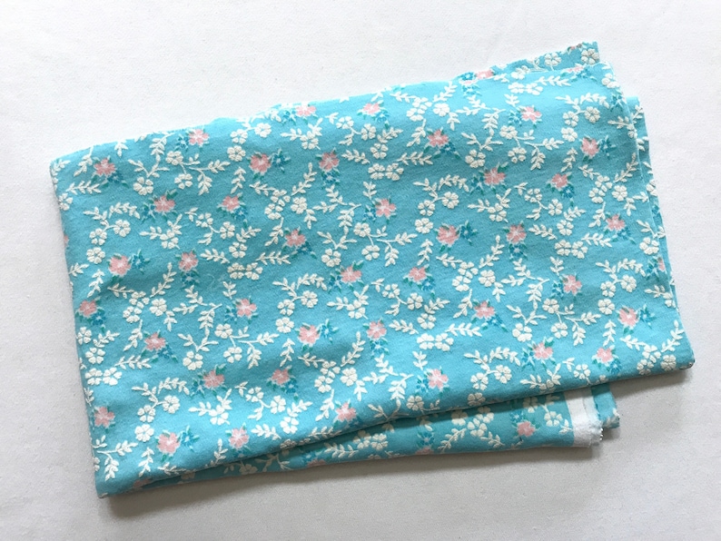 May include: A light blue fabric with a white and pink floral pattern. The fabric is folded in half and has a white border.