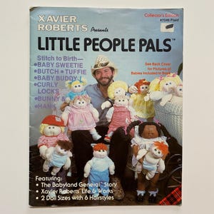 May include: A book cover featuring a man with a group of dolls. The book is titled "Little People Pals" and is a collector's edition of Xavier Roberts' Babyland General dolls. The book includes instructions for making the dolls and features two doll sizes with six hairstyles.