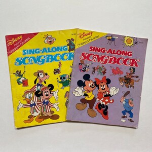 May include: Two vintage Disney "Sing-Along Songbook" books. One book has a yellow cover with Mickey, Donald, and Goofy. The other book has a purple cover with Mickey and Minnie Mouse. Both books feature colourful illustrations of Disney characters.
