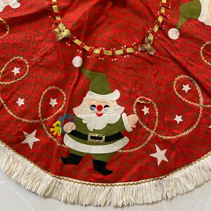 K&ouml;nnte beinhalten: Roter Weihnachtsbaumrock mit sich wiederholendem Blumenmuster und wei&szlig;em Fransen. Der Rock hat eine Filz-Weihnachtsmann-Applikation, goldene Stern- und Girlandenakzente und eine dekorative Bandverzierung.