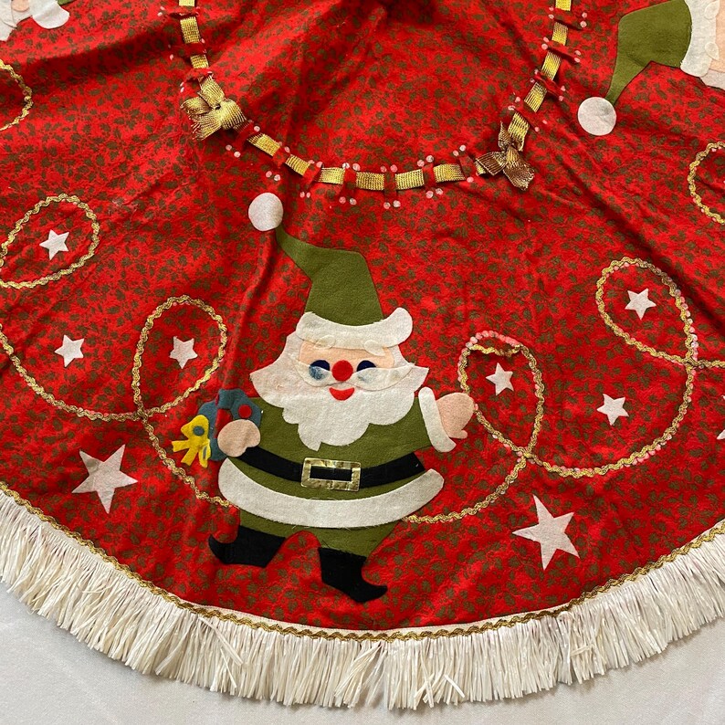 K&ouml;nnte beinhalten: Roter Weihnachtsbaumrock mit sich wiederholendem Blumenmuster und wei&szlig;em Fransen. Der Rock hat eine Filz-Weihnachtsmann-Applikation, goldene Stern- und Girlandenakzente und eine dekorative Bandverzierung.
