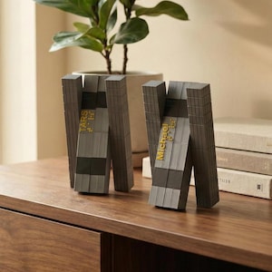 Peut inclure: Deux sculptures grises abstraites avec des lettres dorées et du texte en braille, l'une lisant "TARS" et l'autre "Michael". Les sculptures sont sur une surface en bois, avec une plante en pot en arrière-plan.