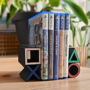 Puede incluir: Un sujetalibros geométrico negro y azul con forma de mando de consola de juegos, que sostiene varias carcasas de juegos de PS4. Las carcasas de los juegos están etiquetadas con títulos como "Sackboy: A Big Adventure" y "Hogwarts Legacy".