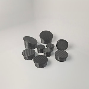 May include: A collection of black, carbon fiber textured plugs of varying sizes. The plugs have a rounded top and a cylindrical base. The image is taken against a plain white background, highlighting the texture and shape of the plugs.